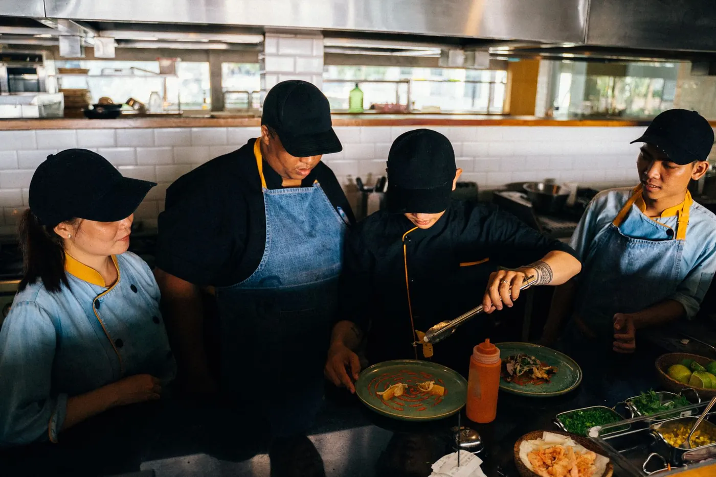 Four chefs in caps and aprons plating dishes together in a kitchen